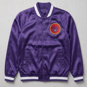 Puede incluir: Una chaqueta bomber de satén morado con cuello, puños y dobladillo acanalados con rayas blancas. La chaqueta tiene un cierre frontal de botones a presión y un parche circular en el pecho izquierdo con un gráfico de dragón estilizado en rojo y amarillo.
