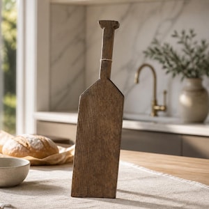 May include: A tall, narrow, dark brown wooden serving board with a bottle-shaped silhouette. The board features a long, slender handle at the top and a wider, rectangular base. The wood grain is visible throughout the surface of the object.