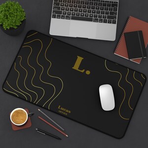 Puede incluir: Alfombrilla de ratón rectangular negra con líneas onduladas doradas y la letra "L." en dorado. El texto "Lucas OFFICE" está impreso en dorado. Un ratón blanco, bolígrafos y una taza de café están sobre el escritorio.