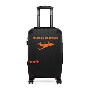Puede incluir: Maleta negra con ruedas y detalles en naranja. La maleta presenta el texto "THE BOSS" y un gráfico de avión naranja. Tres estrellas naranjas están debajo. El asa está extendida.