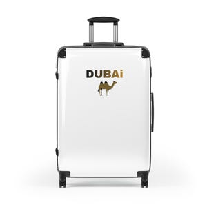 Puede incluir: Maleta rodante blanca con detalles negros y asa retráctil. La maleta presenta la palabra "DUBAI" en dorado y una silueta de camello, ideal para viajar.
