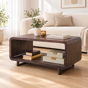 May include: A dark brown wooden coffee table with a rounded rectangular frame and an open lower shelf. The top surface holds a white vase with greenery, a glass candle holder, and a stack of books. The lower shelf contains two books and two stacked rectangular boxes with gold-toned handles.