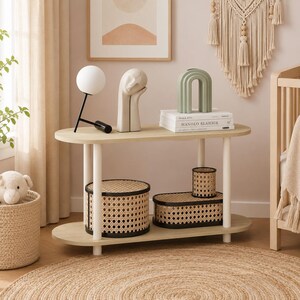 May include: An oval two-tier console table with a light wood finish and white cylindrical legs. The top shelf holds a white globe lamp, a beige abstract face sculpture, a sage green arch vase, and books. The bottom shelf features two rattan storage boxes.
