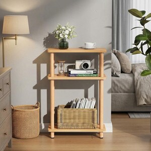 May include: A three-tier light wood shelving unit with rounded corner shelves supported by four cylindrical legs. The top shelf holds a glass vase with white flowers and a white teacup. The middle shelf holds an hourglass, a camera, and books.