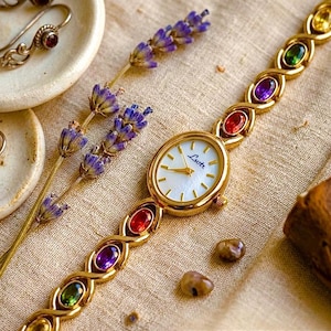 Peut inclure: Montre-bracelet dorée avec un cadran ovale et un bracelet orné de pierres précieuses colorées. Le cadran de la montre porte le mot "Lustre". L'image comprend également des bijoux assortis et des brins de lavande.