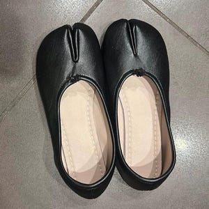 Könnte beinhalten: Ein Paar schwarze Slipper mit einem markanten Zehensteg-Design. Das Obermaterial besteht aus einem glatten, mattschwarzen Material, während das Innere mit einem hellbeigen Stoff ausgekleidet ist. Die Schuhe haben eine flache Silhouette und Form.