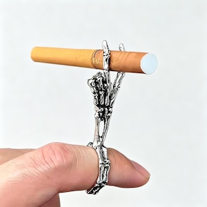 May include: A silver-toned metal ring shaped like a skeletal hand with extended fingers designed to hold a cigarette. The detailed bone structure of the hand forms the band of the ring, with the fingers positioned upright to secure the object.