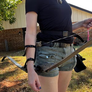 May include: A black recurve bow with a dark wood grain handle and a twisted string, accompanied by a black arm guard with two adjustable straps, a black belt with a plastic buckle, and a black quiver attached to the waist for archery equipment.