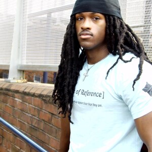 May include: A light blue short-sleeve t-shirt featuring the text "Frame of Reference" printed on the chest, with a small dark star-shaped emblem on the left sleeve. The shirt is worn with a black skull cap and a silver cross necklace.