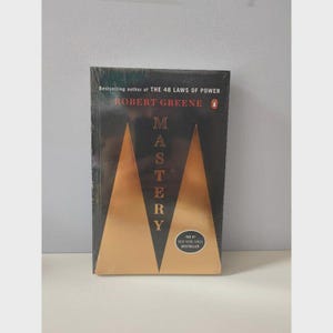 May include: A hardcover book titled "Mastery" by Robert Greene. The cover is black with two gold geometric shapes and the title in gold. The book is a New York Times bestseller.