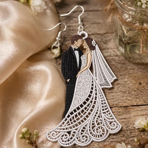 Könnte beinhalten: Gestickte Hochzeitsohrringe mit einem Brautpaar in zärtlicher Umarmung. Das Kleid der Braut ist weiß mit aufwendigen Spitzendetails, und der Bräutigam trägt einen schwarzen Smoking. Die Ohrringe hängen an silbernen Haken.