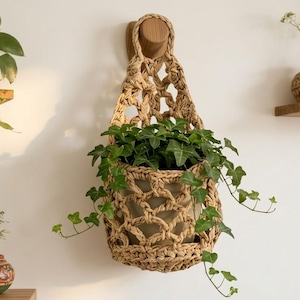 Può includere: Un portapiante in macramè intrecciato con un supporto a parete in legno, che contiene una pianta in vaso con edera verde. Il supporto in fibra naturale è sospeso contro un muro bianco, con altre piante in vaso su mensole.