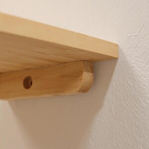 Pu&ograve; includere: Primo piano di una mensola in legno chiaro fissata a un muro bianco. La mensola ha un design semplice e minimalista con un supporto arrotondato e un foro preforato. La venatura del legno &egrave; visibile.