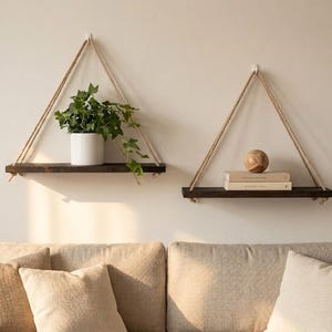 Peut inclure: Deux étagères suspendues en bois brun foncé avec des supports en corde naturelle. L'une contient une plante en pot, l'autre des livres et une sphère en bois. Les étagères sont montées sur un mur de couleur claire, au-dessus d'un canapé beige.