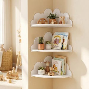 Op de afbeelding: Drie witte hoekplanken met wolkvormige achterwanden gemonteerd aan een muur. De planken bevatten diverse items, waaronder houten speelgoedauto's, kleine vetplantjes in potten, een houten giraffefiguur en diverse kinderboeken met zichtbare titels.