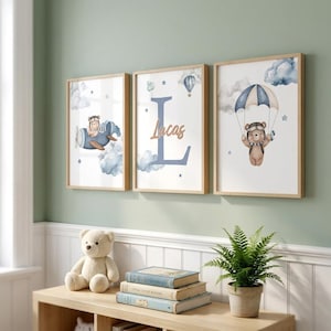 May include: Three framed wall art prints featuring watercolour illustrations of a bear in an aeroplane, a large letter L with the name Lucas, and a bear with a parachute. The prints are displayed in light wood frames against a sage green wall.