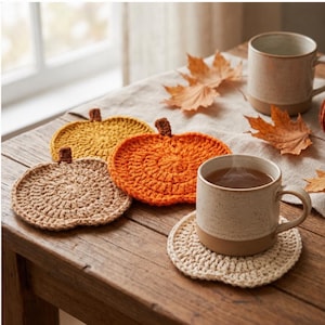 Könnte beinhalten: Ein Set aus vier runden, gehäkelten Untersetzern in Kürbisform in den Farben Beige, Senfgelb und Orange, jeweils mit einem kleinen braunen Stiel. Ein Untersetzer trägt eine gesprenkelte Keramiktasse mit einem Heißgetränk.