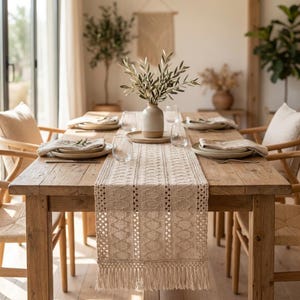 Peut inclure: Un chemin de table en macramé beige avec un motif géométrique et des bords à franges, centré sur une table à manger en bois rustique. La table est dressée avec des assiettes en céramique, des serviettes en lin, des verres et un vase.