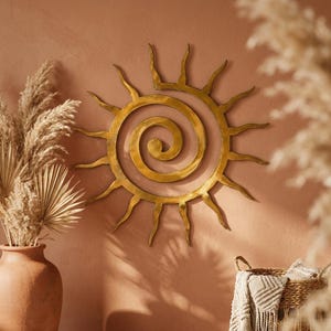 May include: A circular gold-toned metal wall art piece featuring a central spiral design surrounded by radiating, wavy sun-like rays. The object has a smooth, reflective finish and is mounted on a solid, flat surface, creating a distinct sunburst shape.
