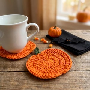 Könnte beinhalten: Eine weiße Keramiktasse steht auf einem runden, orangefarbenen, gehäkelten Kürbis-Untersetzer. Daneben liegen ein weiterer gehäkelter Kürbis-Untersetzer, ein kleiner orangefarbener Kürbis, eine schwarze Stoffserviette mit Fledermaus und Zuckermais.