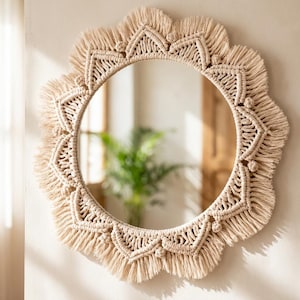 May include: A circular wall mirror framed by intricate off-white macrame cord work. The frame features a repeating geometric pattern of knotted triangles and dense, fringed edges that create a sunburst effect around the central glass.