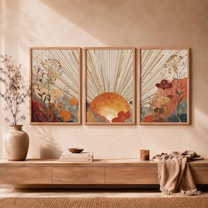 Peut inclure: Un ensemble de trois impressions murales rectangulaires représentant un motif de soleil avec des lignes dorées rayonnantes au-dessus d'un paysage de fleurs sauvages dans des tons orange, rouge et bleu sourd, encadrées de bois clair.