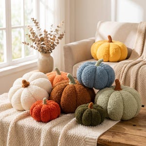 Peut inclure: Une collection de coussins en forme de citrouille, texturés et moelleux, déclinés en plusieurs couleurs : crème, orange, marron, bleu, vert sauge et vert olive foncé. Chaque coussin présente une petite tige distincte et un tissu duveteux.