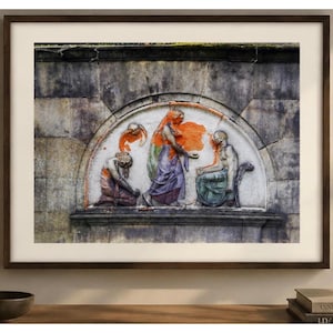 May include: Framed artwork featuring a bas-relief sculpture with figures in shades of orange, purple, and green. The artwork is set against a textured gray background and framed in a dark brown wooden frame. The artwork is displayed on a wooden surface.