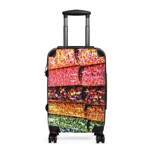 Puede incluir: Una maleta con ruedas, marco, asa y ruedas de color negro. El panel frontal presenta un estampado gráfico vibrante y multicolor de filas de pequeños objetos brillantes similares a caramelos en tonos amarillo, naranja, rojo, morado y verde.