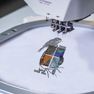 Könnte beinhalten: Eine Stickmaschine, die ein Motiv auf weißen Stoff stickt. Das Design zeigt einen Vogel auf einem Ast mit einer viergeteilten Fenstergrafik, die Landschaftsillustrationen und den Text 'a little bird told me.' enthält.