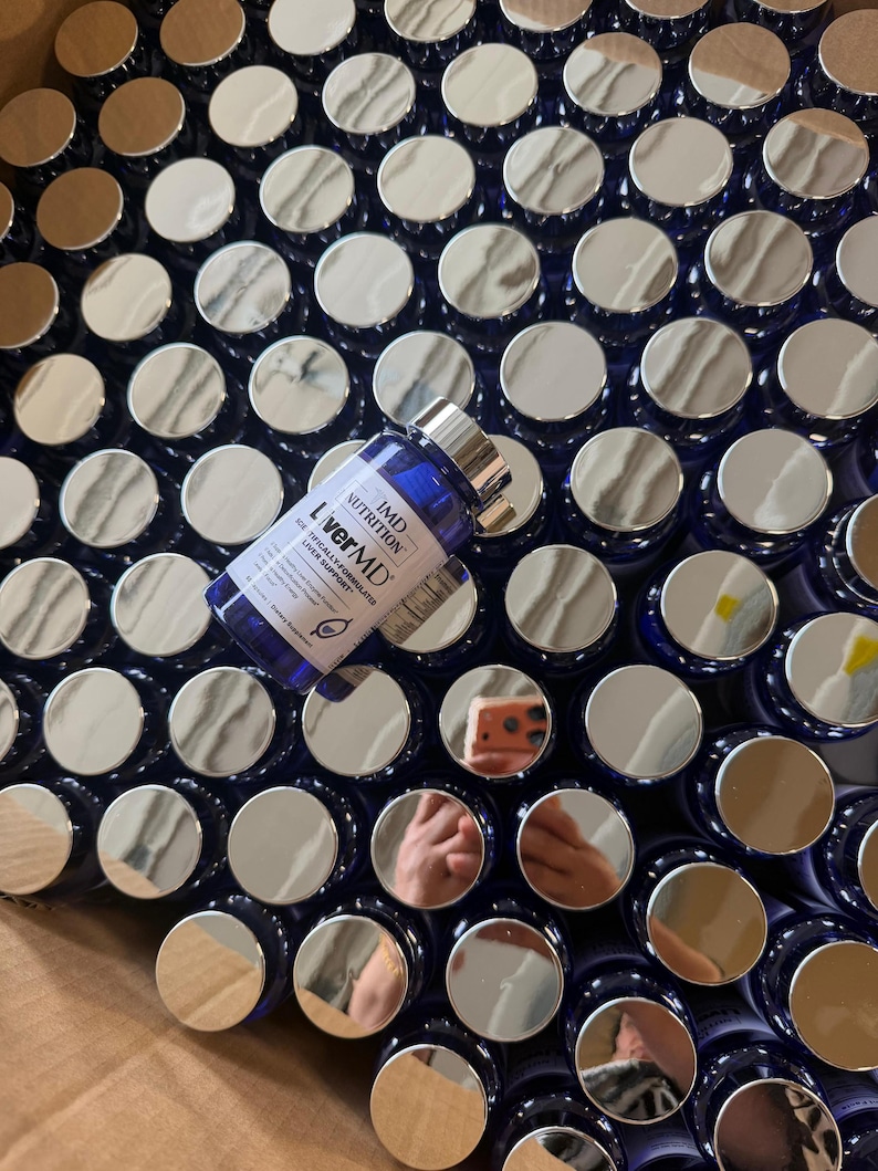 May include: A close-up of numerous blue supplement bottles with silver lids. One bottle is in focus, labelled "LiverMD" and "IMD Nutrition". The bottles are densely packed, indicating a large quantity.
