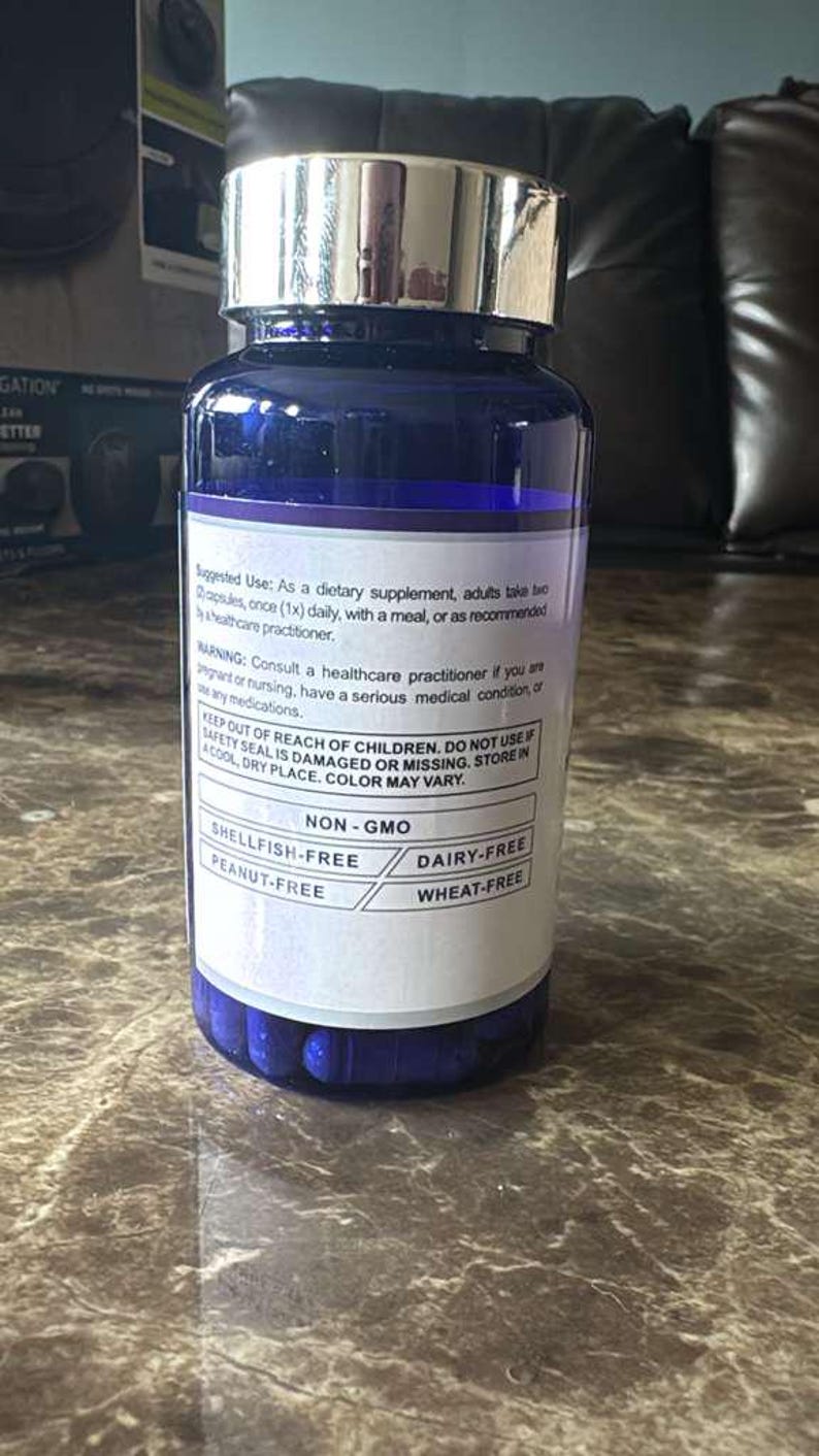 May include: A blue supplement bottle with a silver lid. The label states the product is non-GMO, and free of shellfish, peanuts, dairy, and wheat. The bottle contains blue capsules and is on a marble surface.