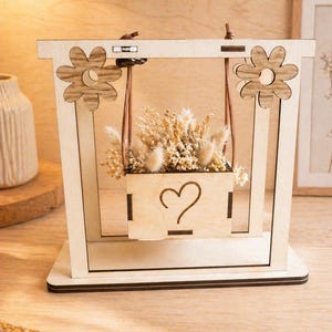 Può includere: Decorazione in legno a forma di altalena con fioriera rettangolare piena di fiori secchi. La fioriera è sospesa da cordoncini di cuoio. Il telaio dell'altalena presenta intagli a forma di fiore e un design a cuore.