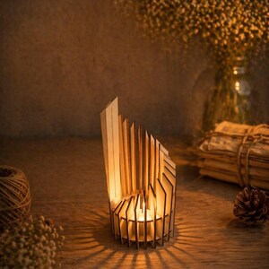 May include: A wooden candle holder with a geometric design. The holder is made of light-coloured wood and has vertical slats that create a pattern. A lit candle is inside, casting shadows. The overall theme is home decor.