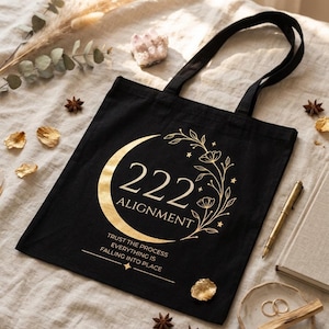 Pode incluir: Uma bolsa tote preta com um gr&aacute;fico dourado de uma lua crescente, detalhes florais e o texto 222 ALIGNMENT. Abaixo do gr&aacute;fico, o texto diz TRUST THE PROCESS EVERYTHING IS FALLING INTO PLACE. A bolsa possui duas al&ccedil;as pretas.