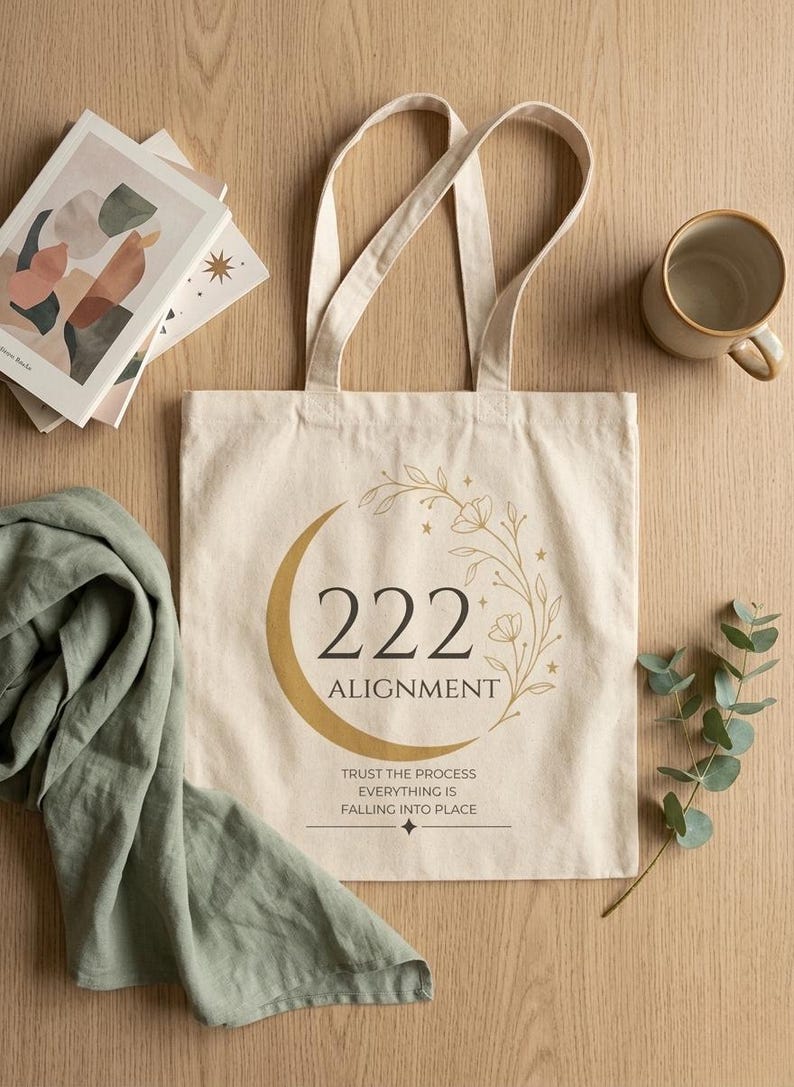 Pode incluir: Uma sacola de lona na cor creme com um gr&aacute;fico de lua crescente dourada, um ramo floral e o texto 222 ALIGNMENT. Abaixo do texto, l&ecirc;-se TRUST THE PROCESS EVERYTHING IS FALLING INTO PLACE com um pequeno s&iacute;mbolo de estrela na parte inferior.