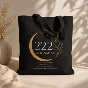 Pode incluir: Uma bolsa tote preta com um gr&aacute;fico dourado de uma lua crescente, vinhas florais e estrelas. O texto diz 222 ALIGNMENT, seguido pela frase TRUST THE PROCESS EVERYTHING IS FALLING INTO PLACE, centralizado com um pequeno &iacute;cone de estrela.