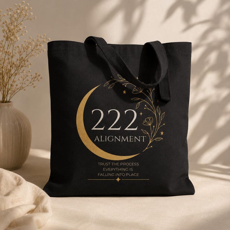 Pode incluir: Uma bolsa tote preta com um gr&aacute;fico dourado de uma lua crescente, vinhas florais e estrelas. O texto diz 222 ALIGNMENT, seguido pela frase TRUST THE PROCESS EVERYTHING IS FALLING INTO PLACE, centralizado com um pequeno &iacute;cone de estrela.