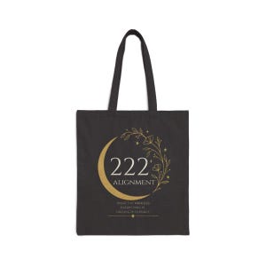 Pode incluir: Uma sacola preta com um gr&aacute;fico dourado de uma lua crescente e vinhas florais. O texto diz 222 ALIGNMENT, seguido pela frase TRUST THE PROCESS EVERYTHING IS FALLING INTO PLACE, com pequenos detalhes de estrelas e linhas horizontais.