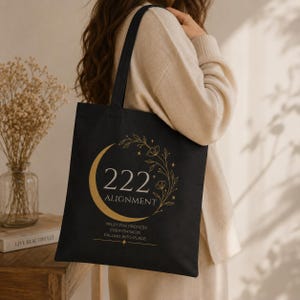 Pode incluir: Uma bolsa tote preta com um gr&aacute;fico dourado de uma lua crescente, vinhas florais e estrelas. O texto diz 222 ALIGNMENT, TRUST THE PROCESS, EVERYTHING IS FALLING INTO PLACE. A bolsa tem duas al&ccedil;as de ombro e um design retangular e plano.