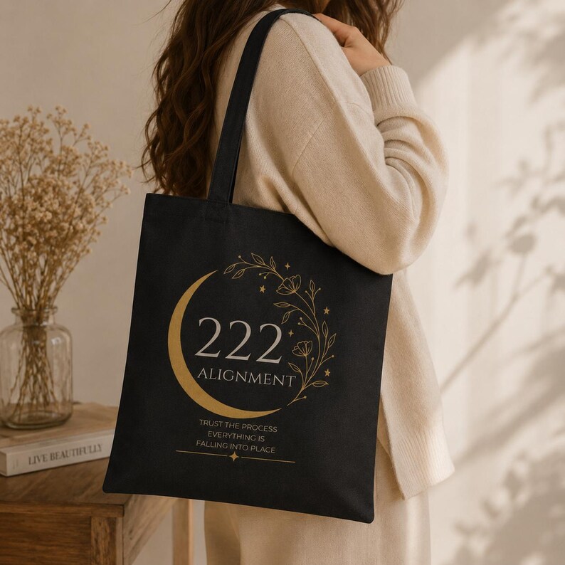 Pode incluir: Uma bolsa tote preta com um gr&aacute;fico dourado de uma lua crescente, vinhas florais e estrelas. O texto diz 222 ALIGNMENT, TRUST THE PROCESS, EVERYTHING IS FALLING INTO PLACE. A bolsa tem duas al&ccedil;as de ombro e um design retangular e plano.