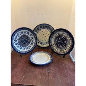 May include: Four ceramic plates with blue and white floral patterns. The plates are displayed on black metal stands and a wooden table. The plates have various designs, including floral and dot patterns. The plates are of different sizes and shapes.