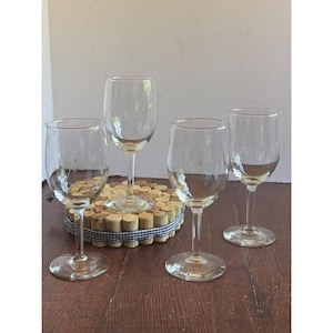 Op de afbeelding: Vier heldere wijnglazen met lange stelen worden getoond. De glazen zijn leeg en van glas. Op de achtergrond een ronde onderzetter van kurken en blauw-wit geruit lint.