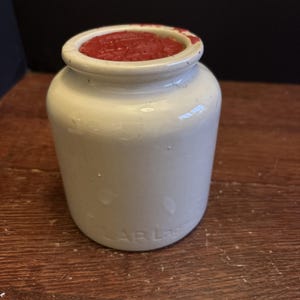 May include: A small, off-white ceramic jar with a wide opening filled with a red substance. The jar has a cylindrical shape with a slightly flared rim. The jar is sitting on a wooden surface.