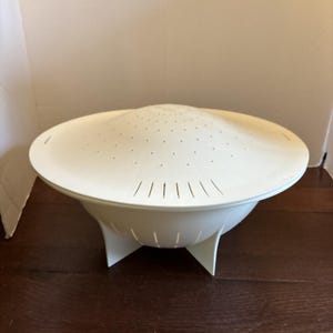 May include: A white plastic colander with a domed lid featuring small circular perforations at the center and elongated vertical slots along the perimeter. The bowl-shaped base is supported by three wide, flat feet extending from the bottom.