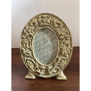 May include: An oval-shaped cream-colored picture frame featuring a raised relief design of flowers and leaves. The center of the frame contains a paper insert printed with the repeating text JOY LEA. The frame stands on two small feet.