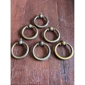 May include: Six antique brass ring pulls with a circular design and a decorative knob. The pulls are arranged on a wooden surface, showcasing their aged patina and classic style. These hardware pieces are ideal for furniture restoration or adding vintage charm.