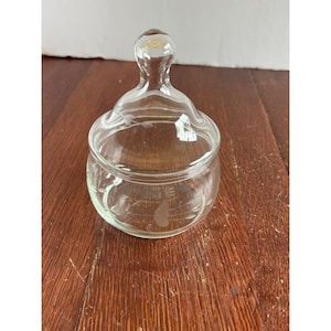 May include: A clear glass candy dish with a matching lid featuring a rounded knob handle. The bowl and lid are decorated with a delicate, etched floral pattern consisting of thin stems and small leaves encircling the exterior of the glass.