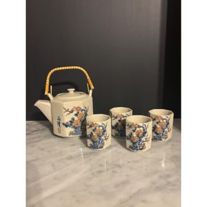 Puede incluir: Un juego de té de cerámica que incluye una tetera octogonal con asa de bambú y cuatro tazas octogonales a juego. Las piezas de color blanco roto están decoradas con motivos florales azules y naranjas y caracteres chinos rojos.