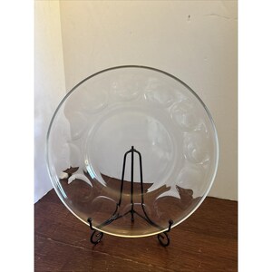 May include: A clear glass plate with a decorative pattern of circles around the rim. The plate is displayed on a black metal stand, set against a neutral background. The plate is round and appears to be made of glass.